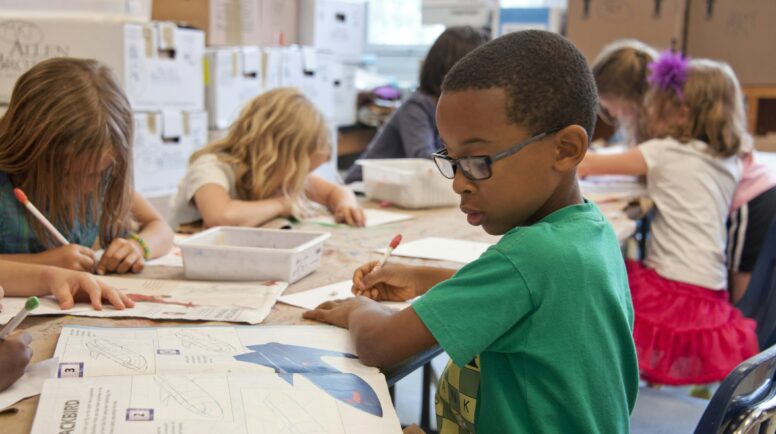 boy in green sweater writing on white paper in a classroom with other children