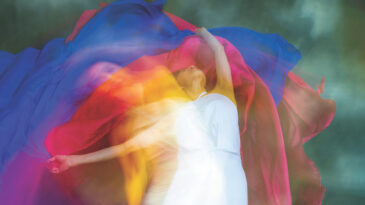 Mystical, blurred image of a female professional dancer in a white dress, dancing with floating, red and blue, gauzy fabrics in a studio, shot against a gray background.