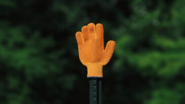 A small orange glove propped up on a fence poll with a green, tree background.