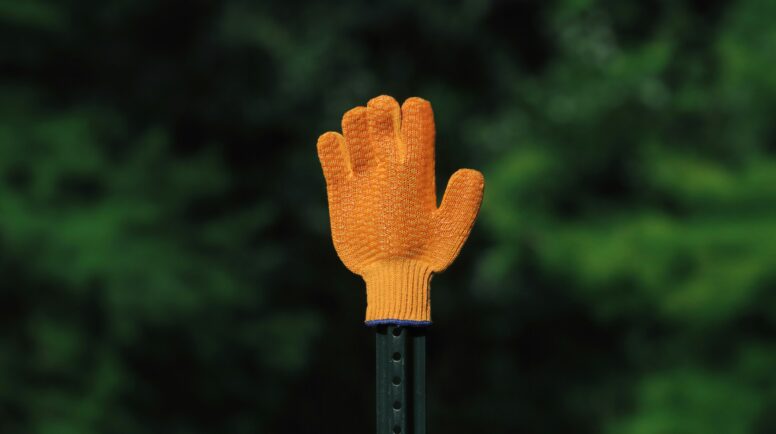 A small orange glove propped up on a fence poll with a green, tree background.