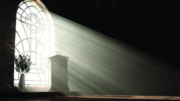 A dark church interior lit by suns rays penetrating through a glass window in the pattern of a crucifix shining on a speech pulpit - 3D render